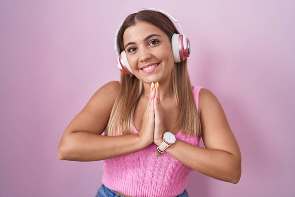 Junge blonde Frau hört Musik über Kopfhörer und betet mit den Händen um Vergebung, lächelt selbstbewusst.  - Foto, Bild