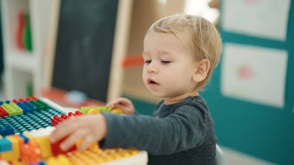 Liebenswertes blondes Kleinkind spielt im Kindergarten mit Bauklötzen am Tisch - Foto, Bild