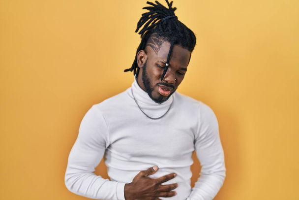 African man with dreadlocks wearing turtleneck sweater over yellow background with hand on stomach because indigestion, painful illness feeling unwell. ache concept.  - Foto, afbeelding