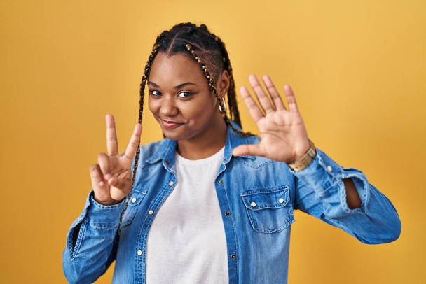 African american woman with braids standing over yellow background showing and pointing up with fingers number seven while smiling confident and happy.  - Foto, Imagem