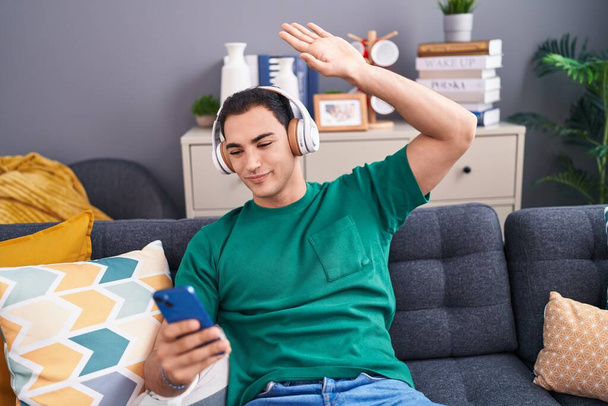 Молодий іспаномовний чоловік танцює і слухає музику, сидячи на дивані вдома
 - Фото, зображення