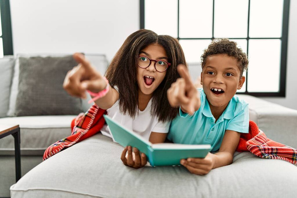 Two siblings lying on the sofa reading a book pointing with finger surprised ahead, open mouth amazed expression, something on the front  - Photo, Image