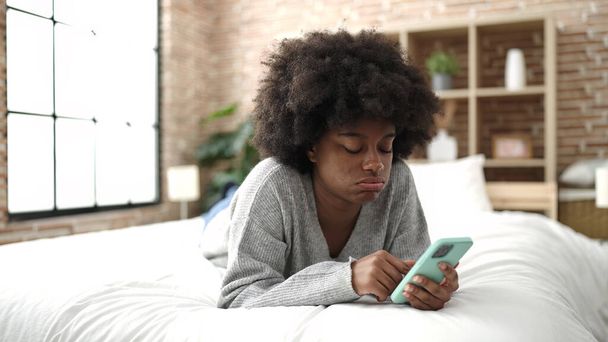 African american woman using smartphone lying on bed at bedroom - Fotó, kép