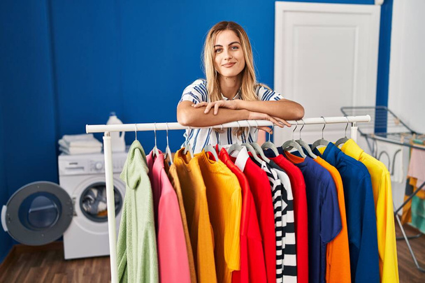 Young blonde woman at laundry room with clean clothes relaxed with serious expression on face. simple and natural looking at the camera.  - Photo, Image