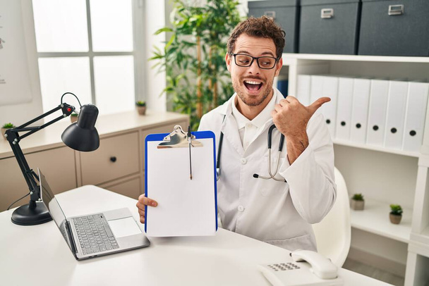 Young hispanic man wearing doctor stethoscope holding clipboard pointing thumb up to the side smiling happy with open mouth  - Фото, изображение