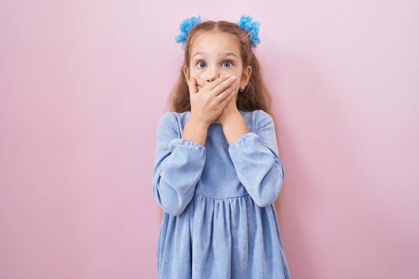 Ein junges kleines Mädchen, das vor rosa Hintergrund steht, schockiert, als es fälschlicherweise den Mund mit den Händen bedeckt. Geheimes Konzept.  - Foto, Bild