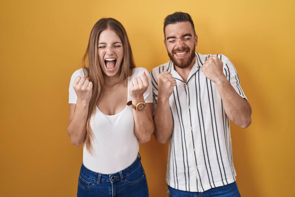 Young couple standing over yellow background excited for success with arms raised and eyes closed celebrating victory smiling. winner concept.  - Fotografie, Obrázek