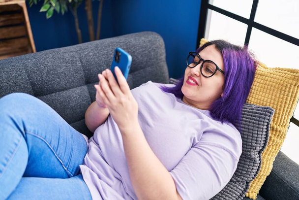 Young beautiful plus size woman using smartphone lying on sofa at home - Fotografie, Obrázek