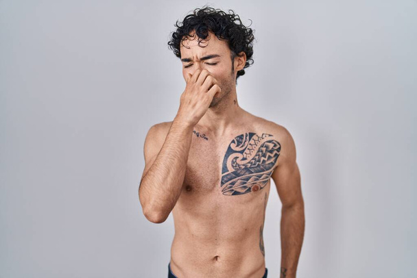 Ισπανόφωνος άνδρας στέκεται γυμνός μυρίζοντας κάτι βρωμερό και αηδιαστικό, ανυπόφορη μυρωδιά, κρατώντας την αναπνοή με τα δάχτυλα στη μύτη. άσχημη μυρωδιά  - Φωτογραφία, εικόνα