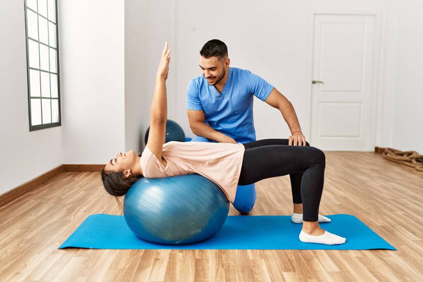 Латинский мужчина и женщина в форме физиотерапевта проходят реабилитационный сеанс, используя фитнес-мяч в реабилитационном центре - Фото, изображение