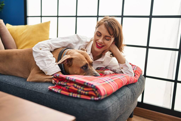 Junge Kaukasierin lächelt selbstbewusst und umarmt Hund, der zu Hause auf Sofa liegt - Foto, Bild