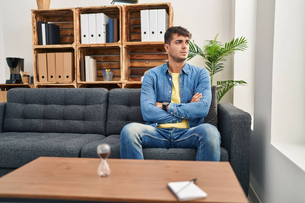 Young hispanic man patient sitting on sofa with serious expression at psychology clinic - Photo, Image