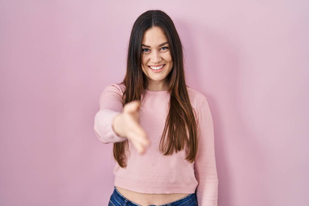 Young brunette woman standing over pink background smiling cheerful offering palm hand giving assistance and acceptance.  - Foto, Imagen