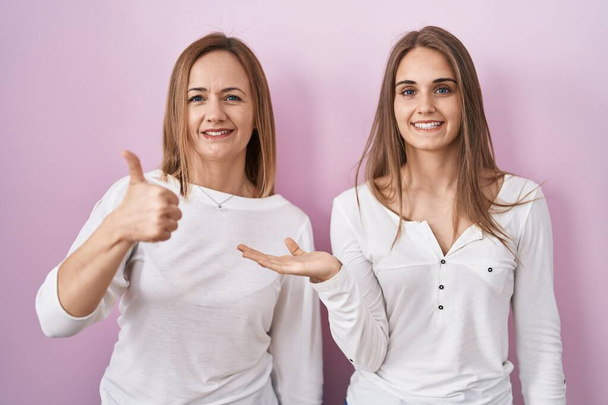Middle age mother and young daughter standing over pink background showing palm hand and doing ok gesture with thumbs up, smiling happy and cheerful  - Foto, afbeelding