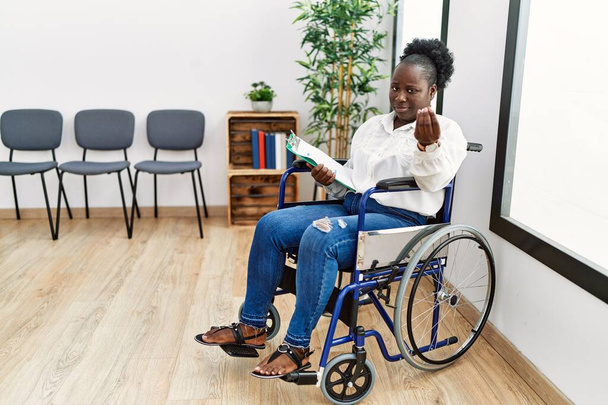 Junge schwarze Frau sitzt im Rollstuhl im Wartezimmer und macht italienische Geste mit selbstbewusstem Ausdruck an Händen und Fingern  - Foto, Bild