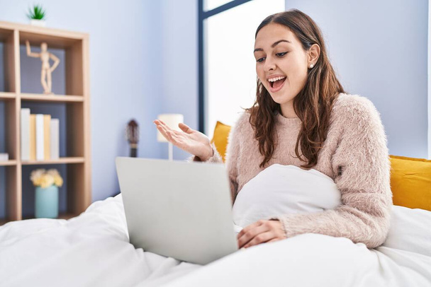Young hispanic woman using computer laptop on the bed celebrating achievement with happy smile and winner expression with raised hand  - Foto, Bild