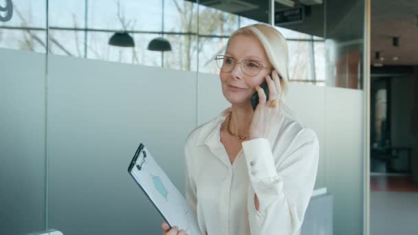 Closeup positive mature business woman 50s in eyeglasses talking by smartphone in office. Smiling businesswoman calling mobile phone in corridor. Portrait of friendly CEO walking in business hallway - Filmagem, Vídeo