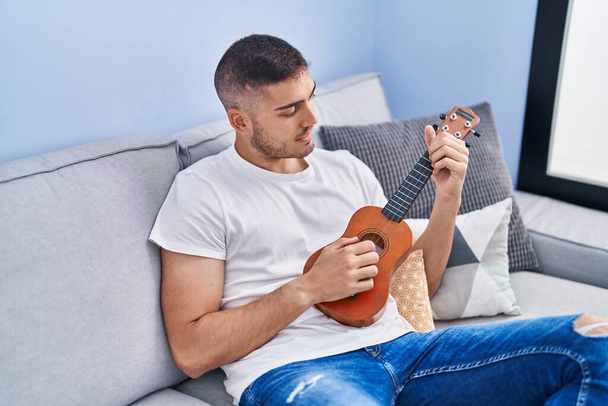 Young hispanic man playing ukelele sitting on sofa at home - Foto, immagini