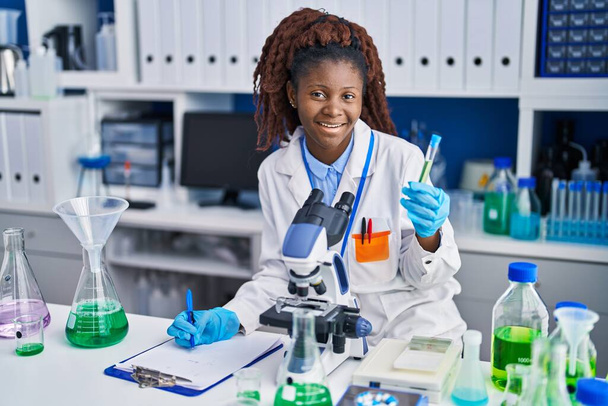 African american woman scientist holding test tube writing report at laboratory - Photo, Image