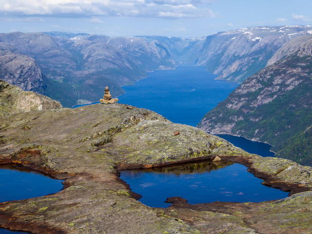 Каменный человек, построенный на краю крутой скалы с видом на Люсефьорд. Каменный человек охраняет окрестности. Живописный вид на фьорд. Несколько луж на вершине утёса, - Фото, изображение