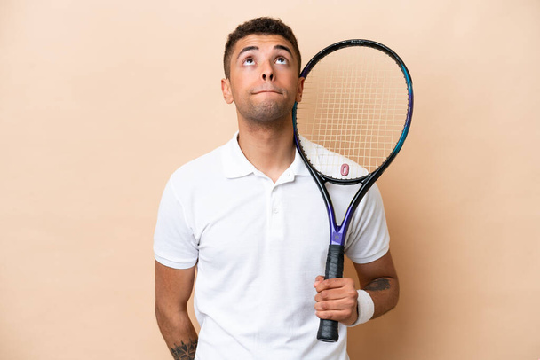 Young brazilian handsome man playing tennis isolated on beige background and looking up - Photo, Image