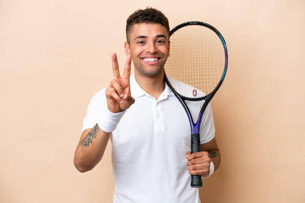 młody brazylijski przystojny mężczyzna gra w tenisa na beżowym tle uśmiechając się i pokazując znak zwycięstwa - Zdjęcie, obraz