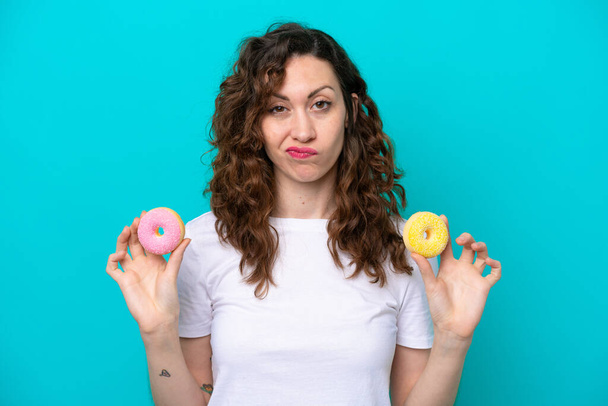 Young caucasian woman isolated on blue background holding donuts with sad expression - Foto, Bild