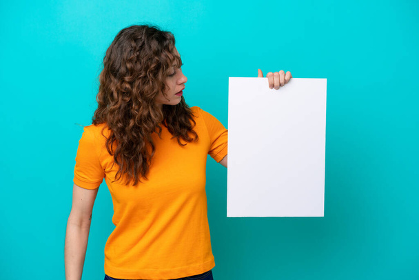 Young caucasian woman isolated on blue background holding an empty placard and looking it - Photo, Image
