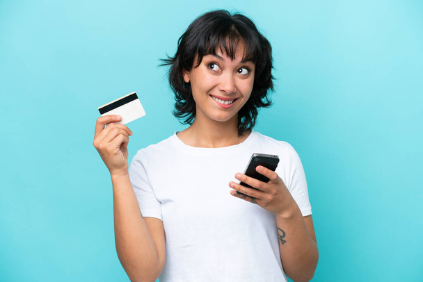 Joven argentina aislada sobre fondo azul comprando con el móvil con tarjeta de crédito mientras piensa - Foto, imagen