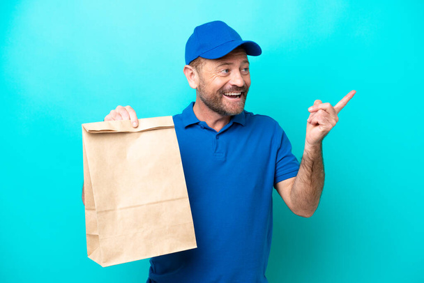 Hombre de mediana edad tomando una bolsa de comida para llevar aislado sobre fondo azul con la intención de darse cuenta de la solución mientras levanta un dedo - Foto, Imagen