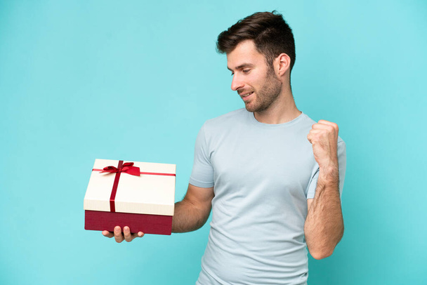 Mavi arka planda elinde bir hediye tutan beyaz bir adam zaferi kutluyor. - Fotoğraf, Görsel