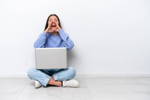 Young caucasian woman with laptop sitting on the floor isolated on white background shouting and announcing something - Photo, Image