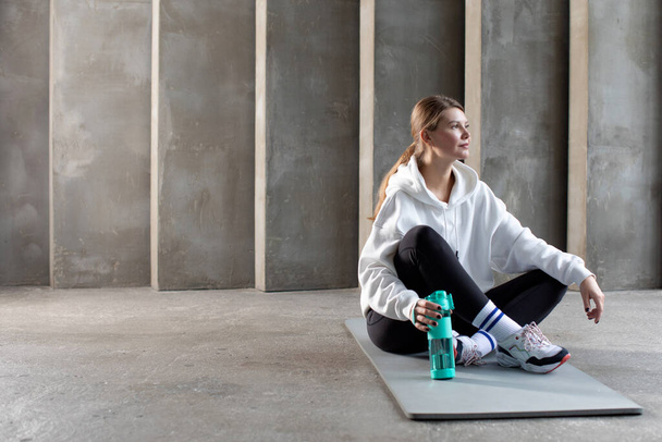 An athlete in sportswear is sitting on a yoga mat with a reusable bottle of water. Drinking balance during workouts. - Foto, imagen