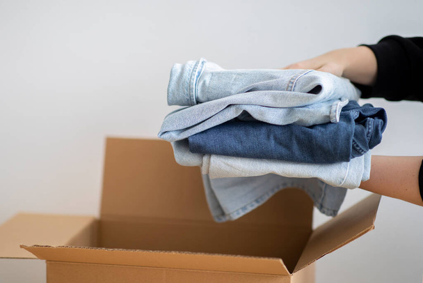Closeup Shot Of Woman Packing Stack Of Clothes In Cardboard Box, Unrecognizable Female Putting Pile Of Jeans To Parcel, Ready To Make Donation Or Sell Used Clothing, Decluttering Home, Cropped Image - Foto, imagen