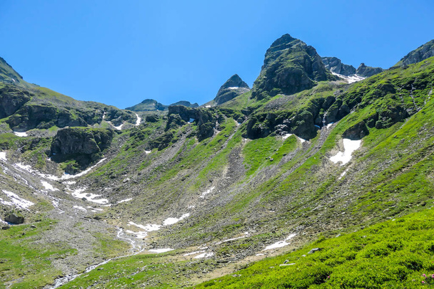 Панорамный вид на Альпы, частично еще покрытые снегом. Весна медленно достигает самых высоких частей гор. Острые вершины, склоны, частично заросшие пышными зелеными растениями. - Фото, изображение