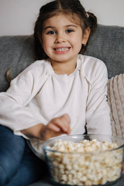 Adorable little girl sits on sofa and watching TV at home. Cute girl eating popcorn. Holiday mood. High quality photo - Fotó, kép