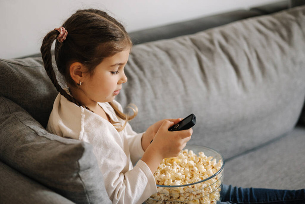 Cute little girl using remote controller of TV and watching cartoons. Four year girl eating popcorn on sofa. High quality photo - Fotó, kép