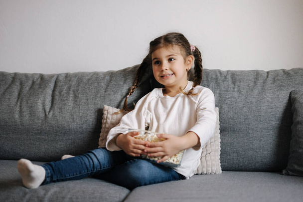 Adorable little girl sits on sofa and watching TV at home. Cute girl eating popcorn. Holiday mood. High quality photo - Fotografie, Obrázek