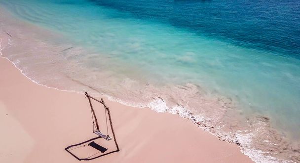 A swing placed on the seashore of Pink Beach, Lombok, Indonesia. The swing has very simple wood construction. Waves gently wash the pillars of it. In the back there are few boats. Drone capture. - Zdjęcie, obraz