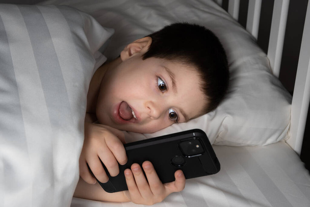 A 5 year old boy plays games on his phone while lying in bed. Gadgets in bed before bedtime in a child. - Photo, Image
