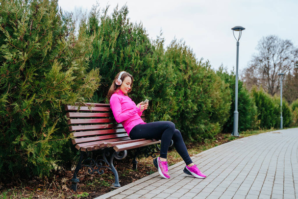 a sportive female athlete takes a moment to rest on a park bench after an intense workout. With her headphones on and smartphone in hand, shes listening to music and catching up on messages - 写真・画像