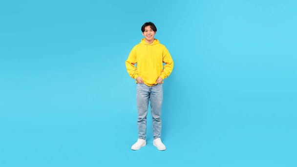 Full Length Shot Of Happy Asian Teen Guy Smiling Looking At Camera Standing Holding Hands In Pockets Over Blue Background. Teenager Boy Wearing Yellow Hoodie And Jeans Posing In Studio. Panorama - Photo, Image