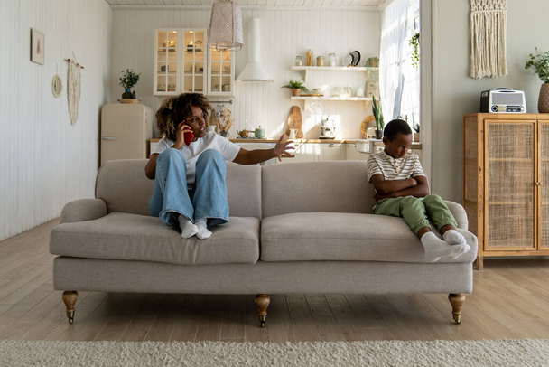 Annoyed angry African American mother sitting with offended upset son on sofa at home, talking on phone, discussing child behavior with spouse. Mom and kid conflict. Parents, children and discipline - Fotografie, Obrázek