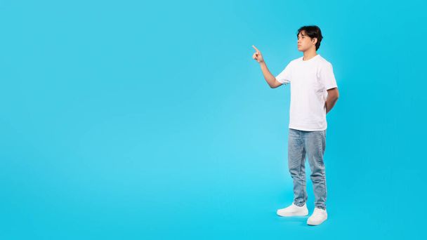 Look There. Serious Asian Teen Guy Pointing Finger Aside Showing Free Space Advertising Your Offer Standing Posing In Studio Over Blue Background. Panorama, Full Length Shot - Fotó, kép