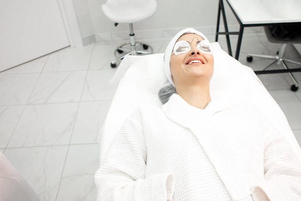 γυναίκα ασθενής βρίσκεται στον καναπέ σε κλινική κοσμετολογίας σε προστατευτικά γυαλιά και χαμόγελα, κορίτσι σε συνεδρία λέιζερ στο αισθητικός - Φωτογραφία, εικόνα