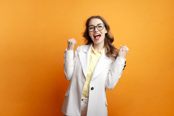 young business girl in glasses and white suit celebrating victory on colored isolated background, woman manager in blazer wins and shows gesture of success - Photo, Image