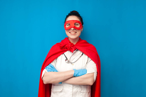 cheerful young girl doctor in uniform in superman costume on blue background, female nurse in superhero mask stands with her arms crossed, healthcare concept - Zdjęcie, obraz