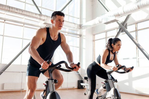 beautiful athletic couple of cyclists train in the fitness room on static bicycle simulator, man trainer and fitness woman go in for sports together on simulators in the morning in bright room - Foto, imagen