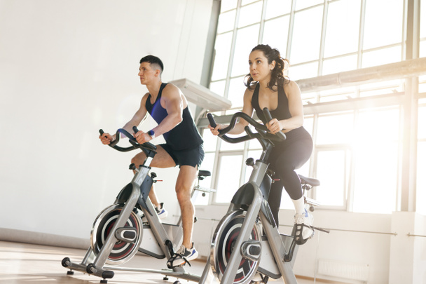 όμορφο αθλητικό ζευγάρι των ποδηλατών τρένο στο γυμναστήριο για στατικό προσομοιωτή ποδήλατο, ο άνθρωπος εκπαιδευτής και fitness γυναίκα πάει για τον αθλητισμό μαζί σε προσομοιωτές το πρωί στο φωτεινό δωμάτιο - Φωτογραφία, εικόνα