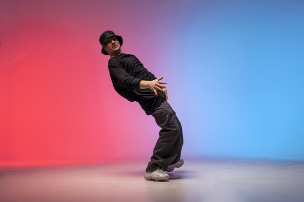 dancer dances hip hop in neon lighting, young guy moves his arms to the sides and dances break dance on red and blue background, elastic choreographer falls to the side, street rap style - Photo, Image